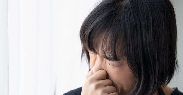 A young woman plugs her nose with her thumb and forefinger as she sneezes in her home. She sits near a white curtain.