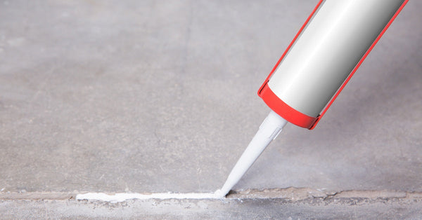 A red caulk gun dispenses a bead of white caulk into an uneven crack in a gray concrete surface to seal it.