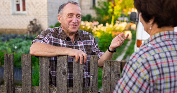 A smiling man has a conversation with his female neighbor across the fence. He rests his arm on the fence.