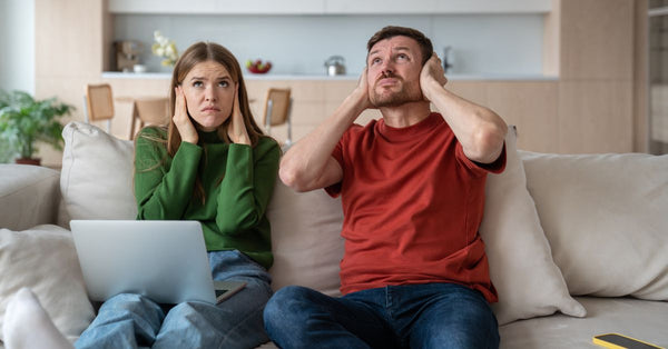 A man and woman sit on the sofa in their living room. They cover their ears as they glare at the ceiling.