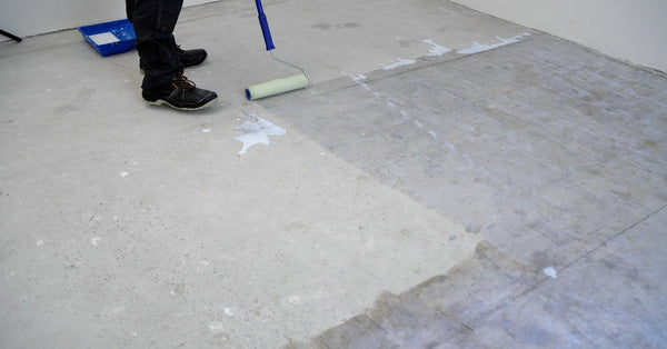 A person uses a paint roller with a long handle to apply a layer of sealant to a concrete basement floor.