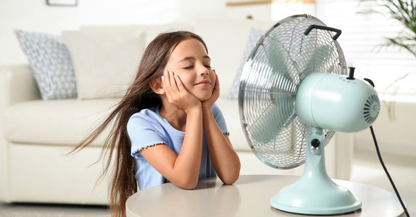 A smiling little girl rests her elbows on a table in front of a fan as she enjoys the cool breeze with her eyes closed.