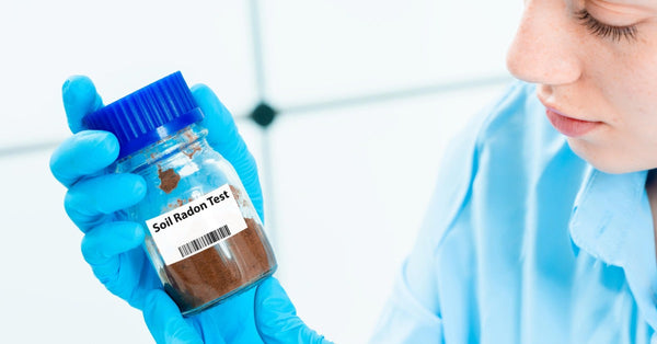 A close-up view shows a woman wearing blue gloves holding a glass jar full of dirt with a label that reads "soil radon test."