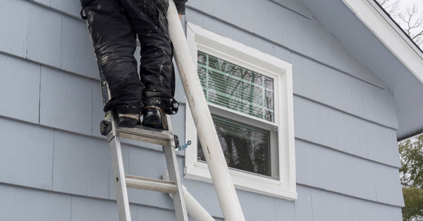 One man is standing on a steel ladder holding a white pipe. Another man stands on the ground with his hands on the ladder.