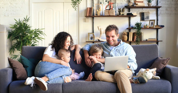 A baby and a toddler sit on a dark blue couch sandwiched between their parents. The whole family is smiling.
