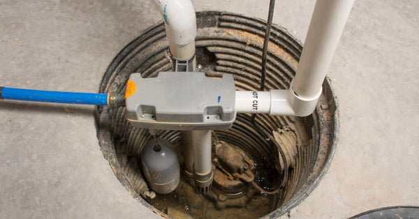 An aerial view shows the inside of a sump pump system, surrounded by concrete. There's a gray pump, PVC pipes, and a blue tube.