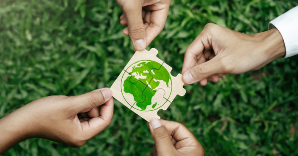 The hands of four individuals each hold puzzle pieces that form a map of earth above a patch of grass.