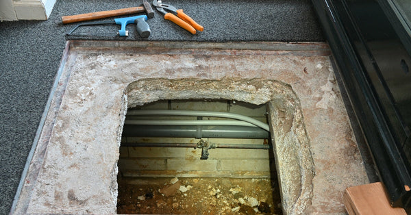 Hand tools sit near an opening in a home's floor that leads to a dirt crawl space lined with utility pipes.