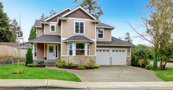 A two-story home with tan siding and a landscaped yard sits on a corner lot in a neighborhood on a sunny day.