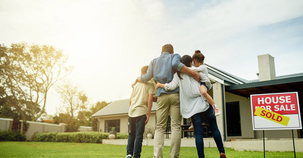 A family with two parents and two kids stand on the front lawn facing a home. A sign reads "House for Sale-Sold."