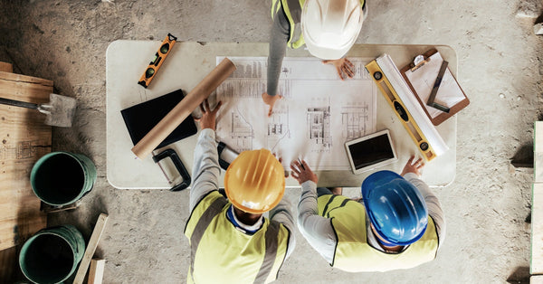 An aerial view shows three people wearing hard hats and yellow safety vests looking at home construction plans.
