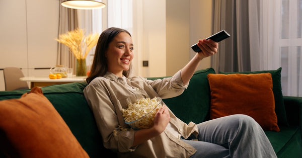 A young woman sits on a green couch holding a bowl of popcorn in one hand and a television remote in the other.