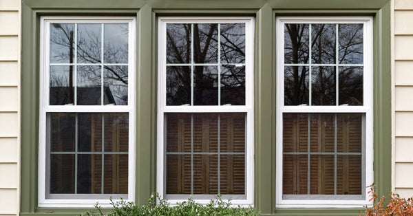 A set of three windows surrounded by olive-green trim nestles into the side of a white-sided home. A bush is in front.