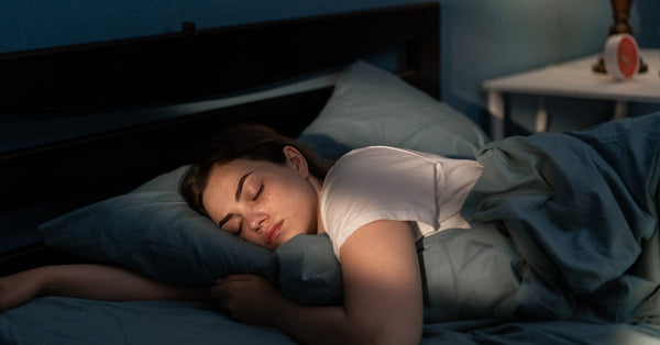 A young woman wearing a white T-shirt is sleeping in a dimly lit room. The bed has dark-colored sheets.