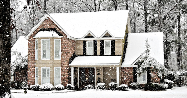 A two-story home features tan siding, brown brick, and black shutters next to the windows. Snow is falling on the home.
