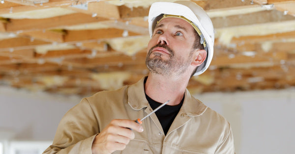 A man wearing a white hard hat and tan jumpsuit holds a flat-head screwdriver, looking at the beams of an exposed ceiling.