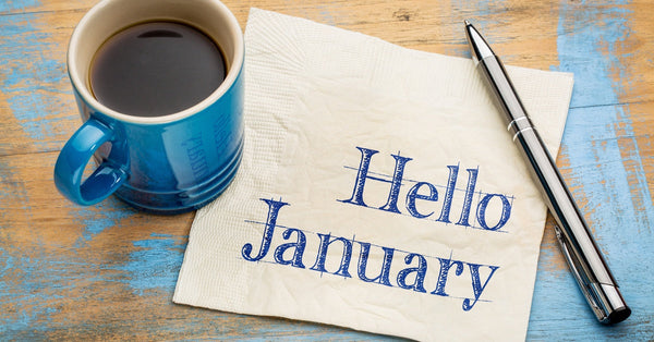 A tan napkin contains the saying "Hello January" written in blue ink. A cup of coffee and a pen are next to the napkin.