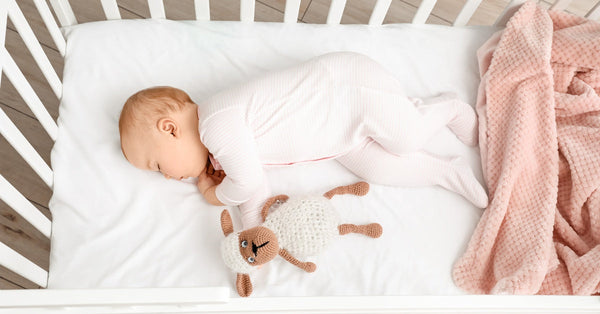 A baby wearing a white sleeper sleeps in a crib with white sheets. A stuffed lamb and a pink blanket are in the crib.