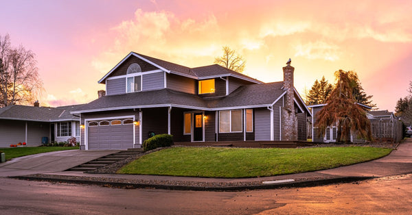 A two-story home with a two-car garage and gray siding sits on a corner lot. The sunset is turning the sky purple and pink.