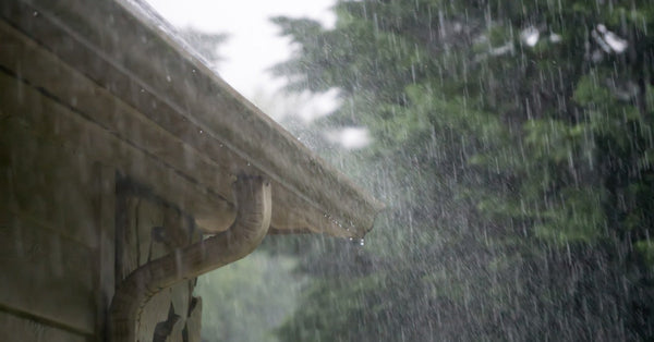 A close-up view shows a heavy rain hitting the gutters, siding, and shingles of a home. A tree is in the background.